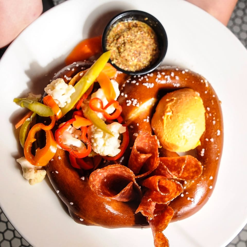 Soft pretzel topped with mustard, cheese, pickled vegetables, and crispy pepperoni on a white plate.