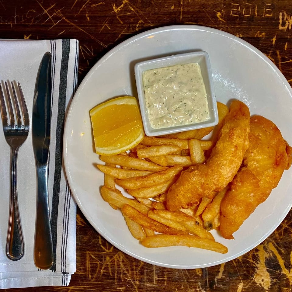 Plate of battered fish with fries, lemon wedges, and tartar sauce beside silverware on a rustic wooden table.