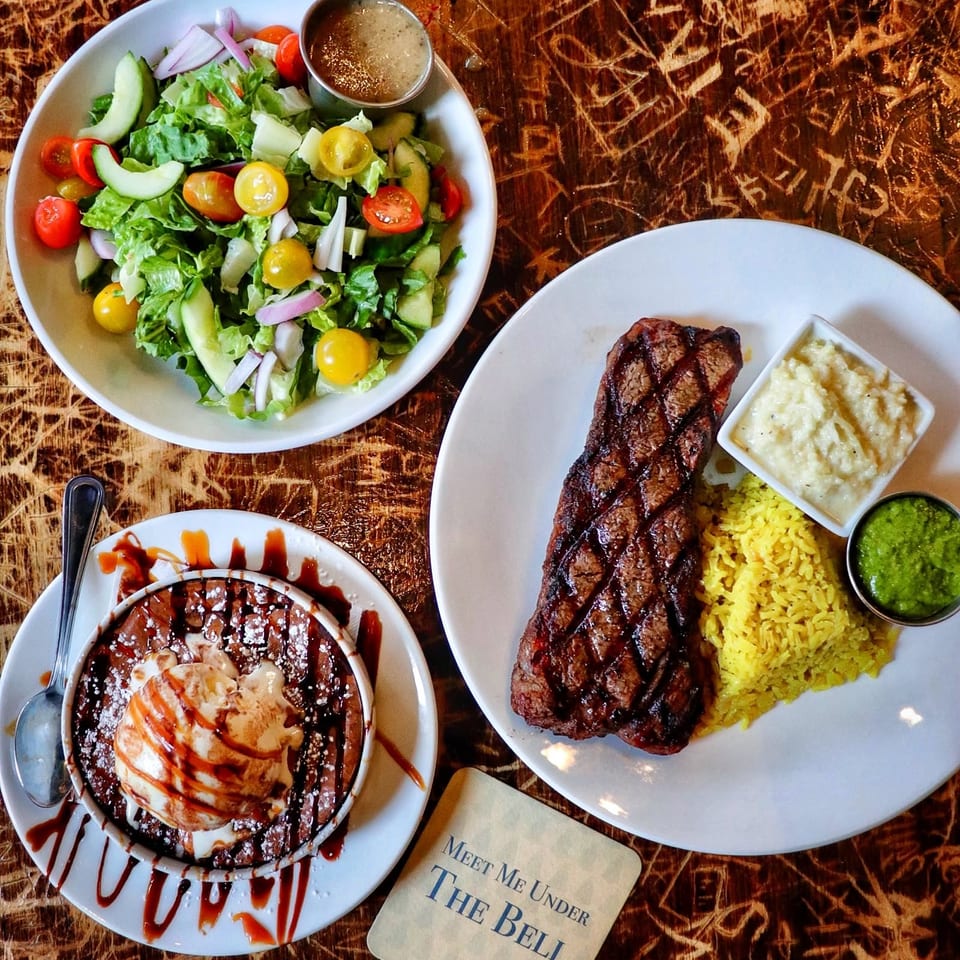 Overhead view of grilled steak with yellow rice, creamy side, salad, and chocolate dessert on a rustic wooden table.