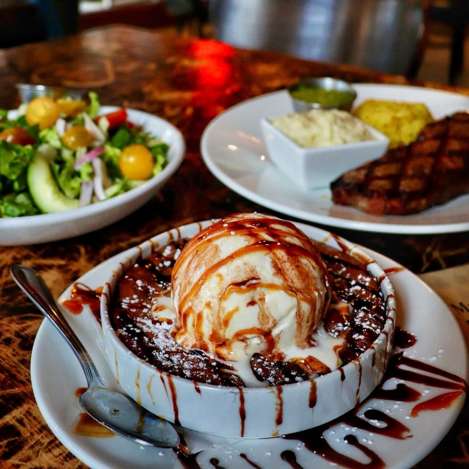 Chocolate brownie topped with vanilla ice cream, chocolate and caramel drizzles, alongside salad and grilled food on a restaurant table.