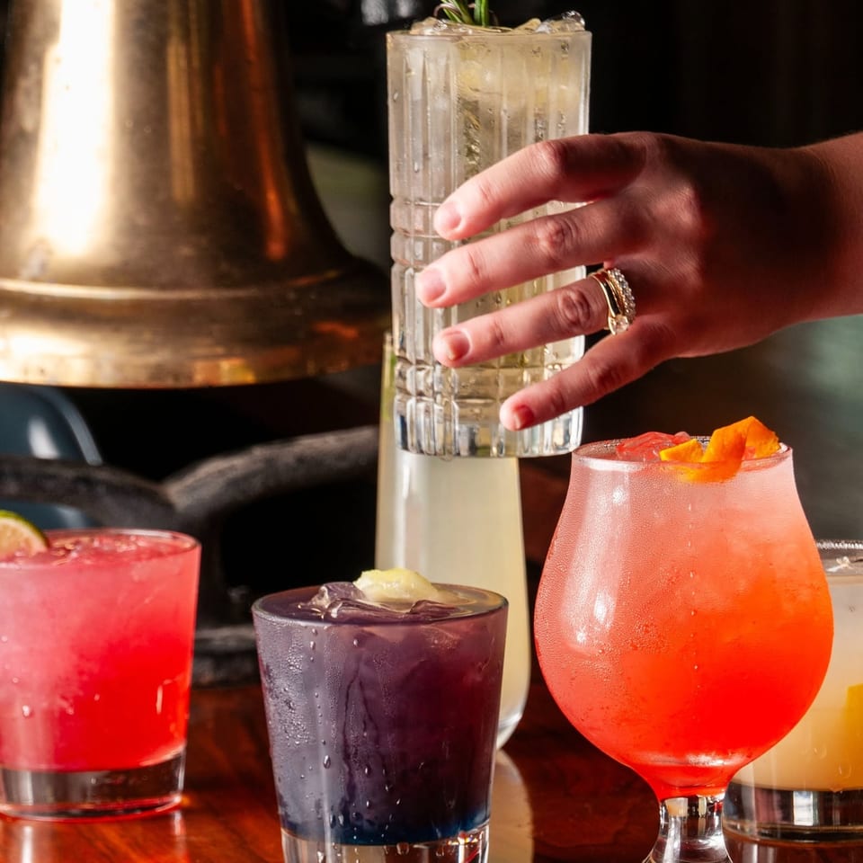Colorful cocktails sit on a polished wooden bar, including a rosemary-garnished drink, while a hand reaches for one.