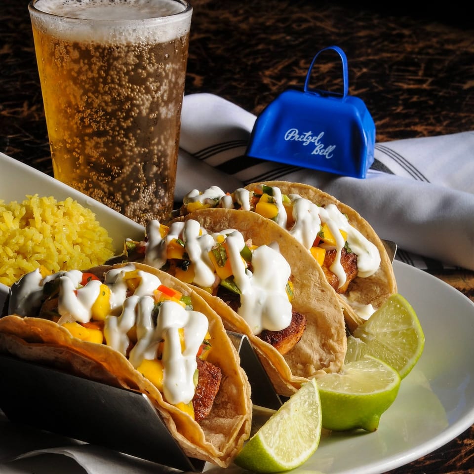 Tacos topped with creamy sauce, diced vegetables, rice, lime wedges, sparkling beer, and a blue pretzel bell on a restaurant table.