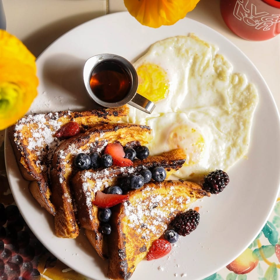 French toast topped with powdered sugar and berries, served with fried eggs, syrup, flowers, and a red coffee mug.
