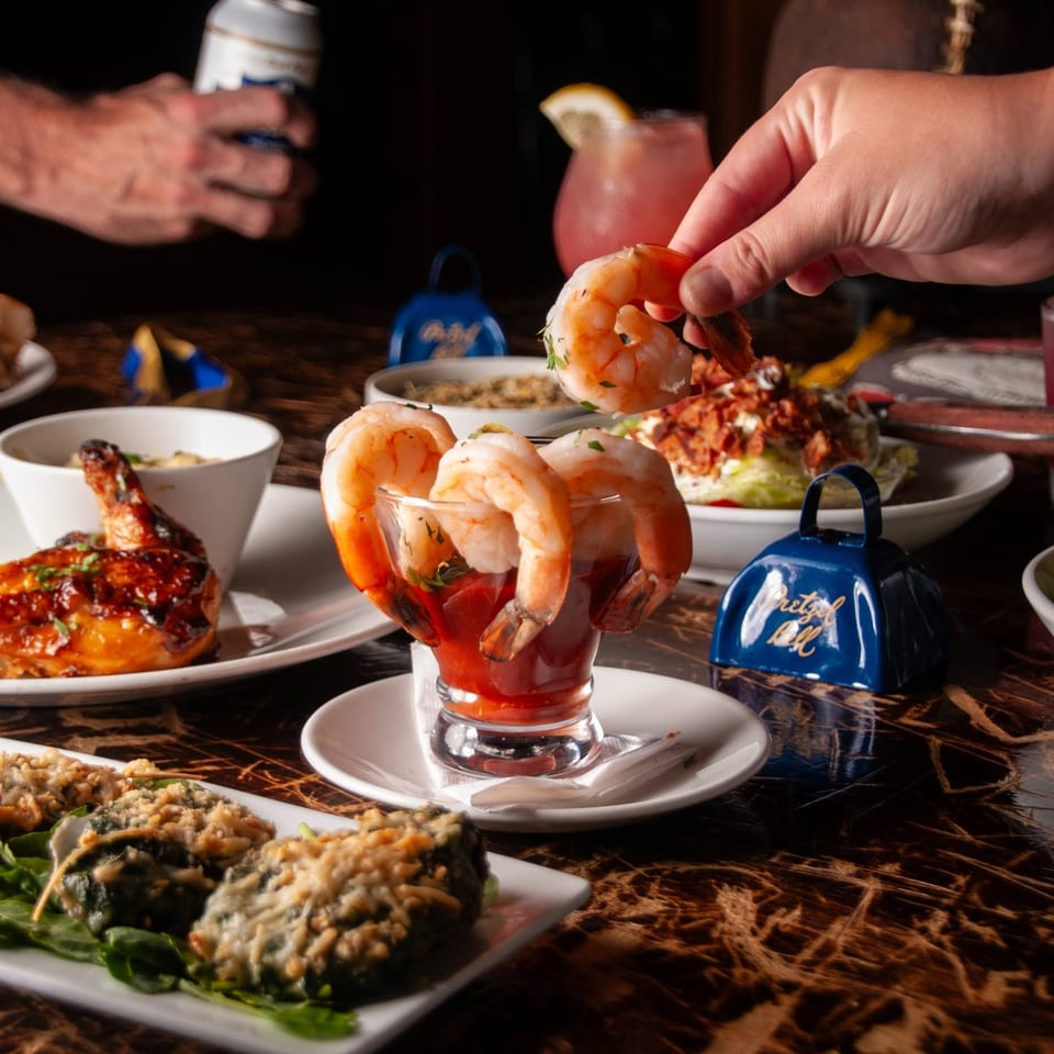 Shrimp cocktail garnished with lemon beside assorted seafood dishes on a restaurant table, with diners’ hands serving food.