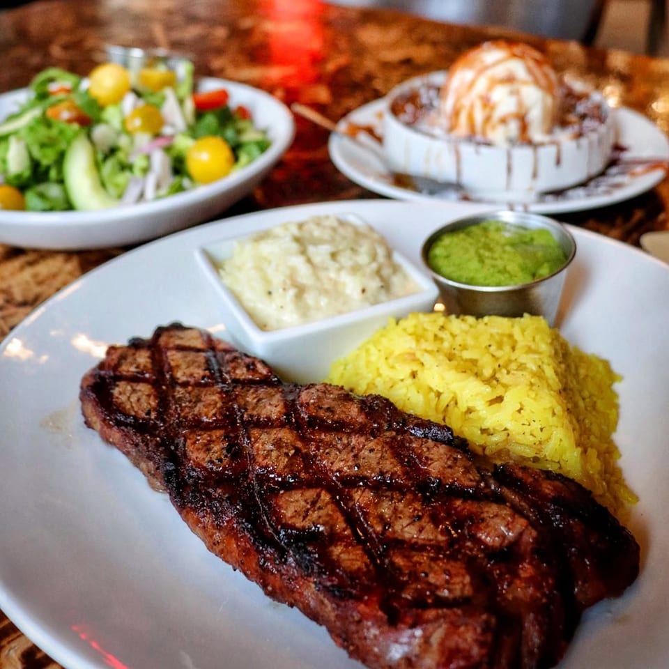 Grilled steak with yellow rice, mashed potatoes, green sauce, salad, and a dessert on a restaurant table.